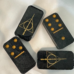 May include: Four black dominoes with gold accents. Two dominoes feature a gold symbol of a wand and triangle. Two dominoes feature gold dots and a gold line.
