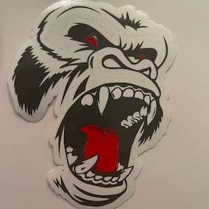May include: A black and white illustration of a gorilla's face with a red tongue sticking out. The gorilla is roaring with its mouth open and teeth showing.