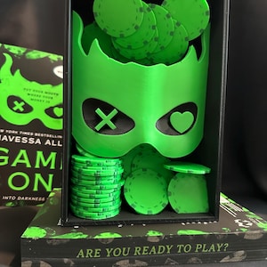 K&ouml;nnte beinhalten: Ein neongr&uuml;ner Pokerchip-Halter mit Maskenmotiv, gef&uuml;llt mit gr&uuml;nen Pokerchips. Der Halter befindet sich in einer schwarzen Box, die auf einem Buch mit dem Text "ARE YOU READY TO PLAY?" ruht. Das Buchcover tr&auml;gt den Text "GAME ON".