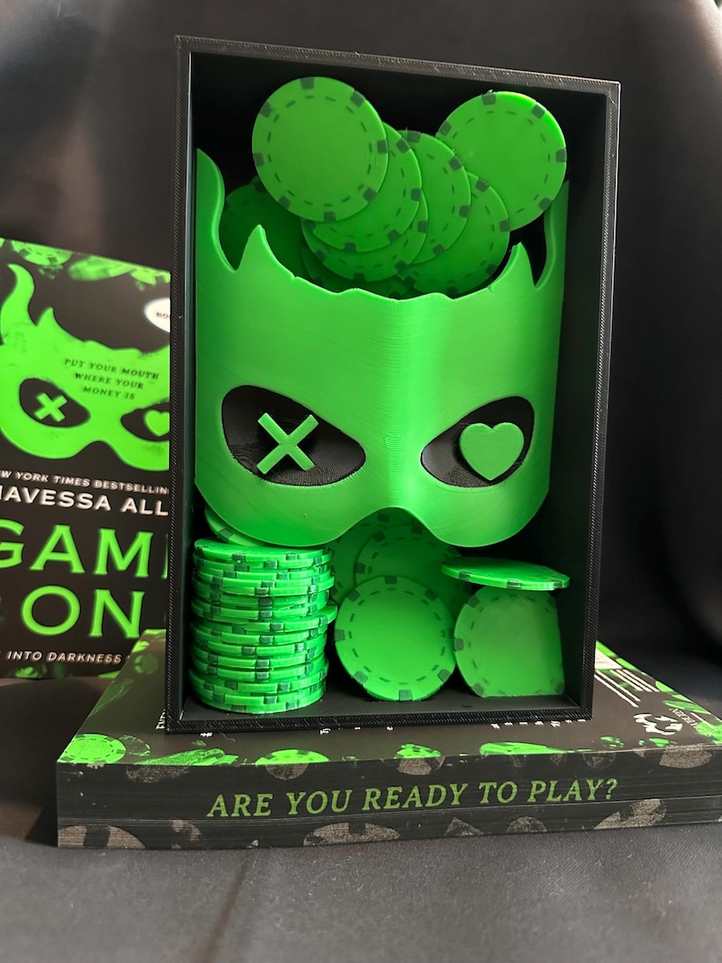 K&ouml;nnte beinhalten: Ein neongr&uuml;ner Pokerchip-Halter mit Maskenmotiv, gef&uuml;llt mit gr&uuml;nen Pokerchips. Der Halter befindet sich in einer schwarzen Box, die auf einem Buch mit dem Text "ARE YOU READY TO PLAY?" ruht. Das Buchcover tr&auml;gt den Text "GAME ON".