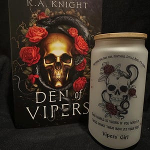 May include: A book titled "Den of Vipers" by K.A. Knight, featuring a skull and snake design, and a frosted glass tumbler with a skull, rose, and snake design. The tumbler has text that reads "Vipers' Girl".