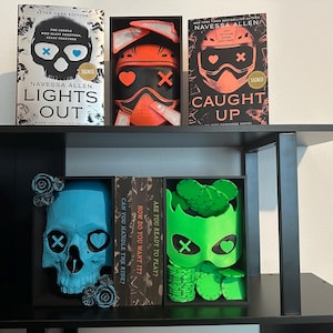 K&ouml;nnte beinhalten: Ein schwarzes B&uuml;cherregal zeigt signierte B&uuml;cher und dekorative Sch&auml;del. Die B&uuml;cher tragen die Titel "Lights Out" und "Caught Up". Die Sch&auml;del sind blau und gr&uuml;n, mit Herz- und Kreuzmotiven. Das Regal ist schwarz.