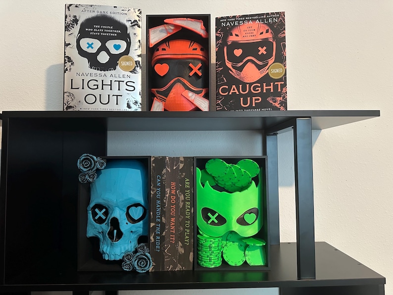 K&ouml;nnte beinhalten: Ein schwarzes B&uuml;cherregal zeigt signierte B&uuml;cher und dekorative Sch&auml;del. Die B&uuml;cher tragen die Titel "Lights Out" und "Caught Up". Die Sch&auml;del sind blau und gr&uuml;n, mit Herz- und Kreuzmotiven. Das Regal ist schwarz.