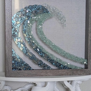 May include: A framed artwork depicting a stylized ocean wave. The wave is crafted from various shades of blue and clear glass pieces, creating a shimmering effect. The artwork is set against a neutral background and framed in a gray wooden frame.