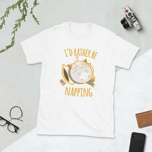 Könnte beinhalten: Weißes T-Shirt mit dem Text "I'd rather be napping" und einer Cartoon-Katze, die auf einem Kissen schläft. Die Katze ist grau und weiß mit orangefarbenen Akzenten. Eine Fernbedienung liegt neben dem Kissen. Das Shirt ist aus weichem Material.