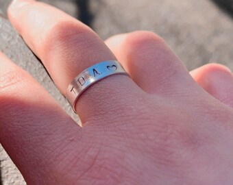 Puede incluir: Un anillo de plata con las letras estampadas "TDA 3" en la banda. El anillo se lleva en un dedo, con un tono de piel claro. El anillo es el foco principal, con un fondo borroso.