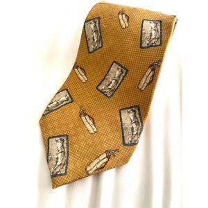 May include: Mustard-colored tie with a repeating pattern of golf bags and golfers in rectangular frames with blue borders. The tie has a textured weave and is displayed against a white background.