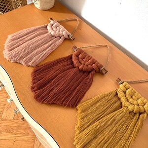 May include: Three macrame wall hangings with fringe in pink, rust, and yellow. Each hanging features a woven circle design and a wooden dowel.