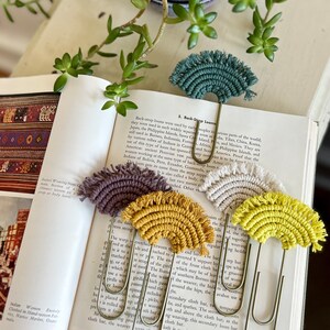 May include: Five colorful rainbow-shaped macrame bookmarks with fringe details. The bookmarks are attached to gold metal paper clips.