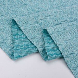 May include: A close-up of a teal blue fabric with a textured, woven pattern. The fabric is folded in half, showing the pattern on both sides.