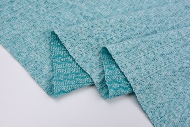 May include: A close-up of a teal blue fabric with a textured, woven pattern. The fabric is folded in half, showing the pattern on both sides.