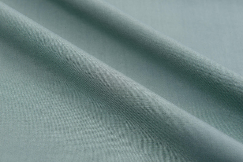 May include: A close-up of a smooth, light blue fabric with a subtle texture. The fabric is folded in a way that creates soft, flowing lines.