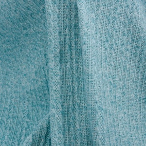 May include: Close-up of a teal blue knitted fabric with a cable knit pattern. The fabric is folded in half, showing the texture of the knit.