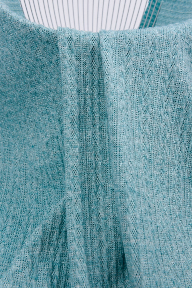 May include: Close-up of a teal blue knitted fabric with a cable knit pattern. The fabric is folded in half, showing the texture of the knit.