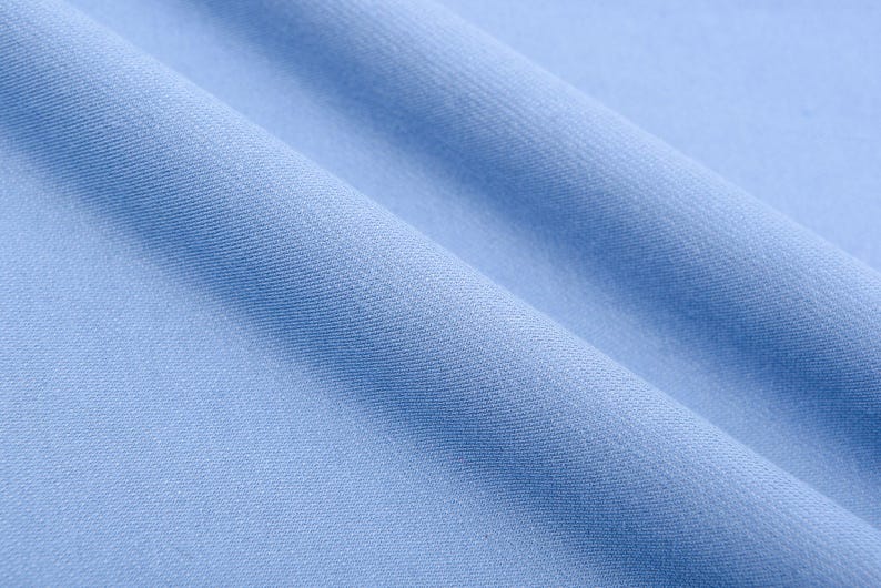 May include: Close-up of light blue fabric with soft folds. The fabric has a smooth texture and a subtle sheen, creating a sense of depth and movement. The color is a pale, calming blue, suitable for various textile applications.