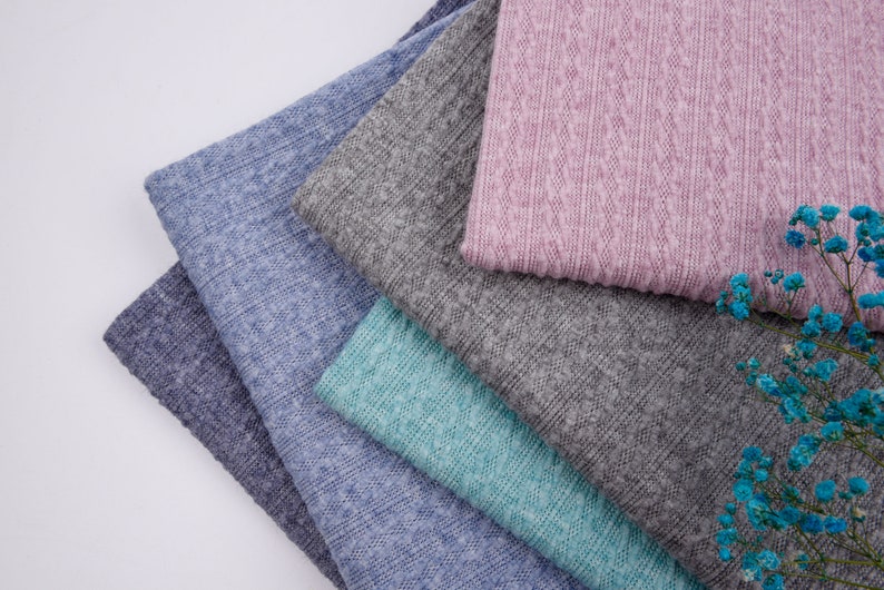 May include: A stack of five fabric swatches in different colors and textures. The top swatch is a light pink with a textured pattern. The other swatches are in shades of blue and green.