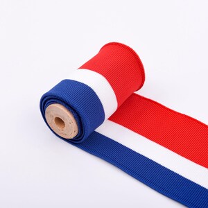May include: A roll of red, white, and blue ribbon. The ribbon is unrolled, revealing the colors in stripes. The roll is made of a ribbed fabric and is on a white background. The ribbon is likely used for crafts or decorations.