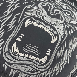 May include: A black and silver embroidered patch of a gorilla's face with a fierce expression. The gorilla's teeth are bared and its eyes are wide open.