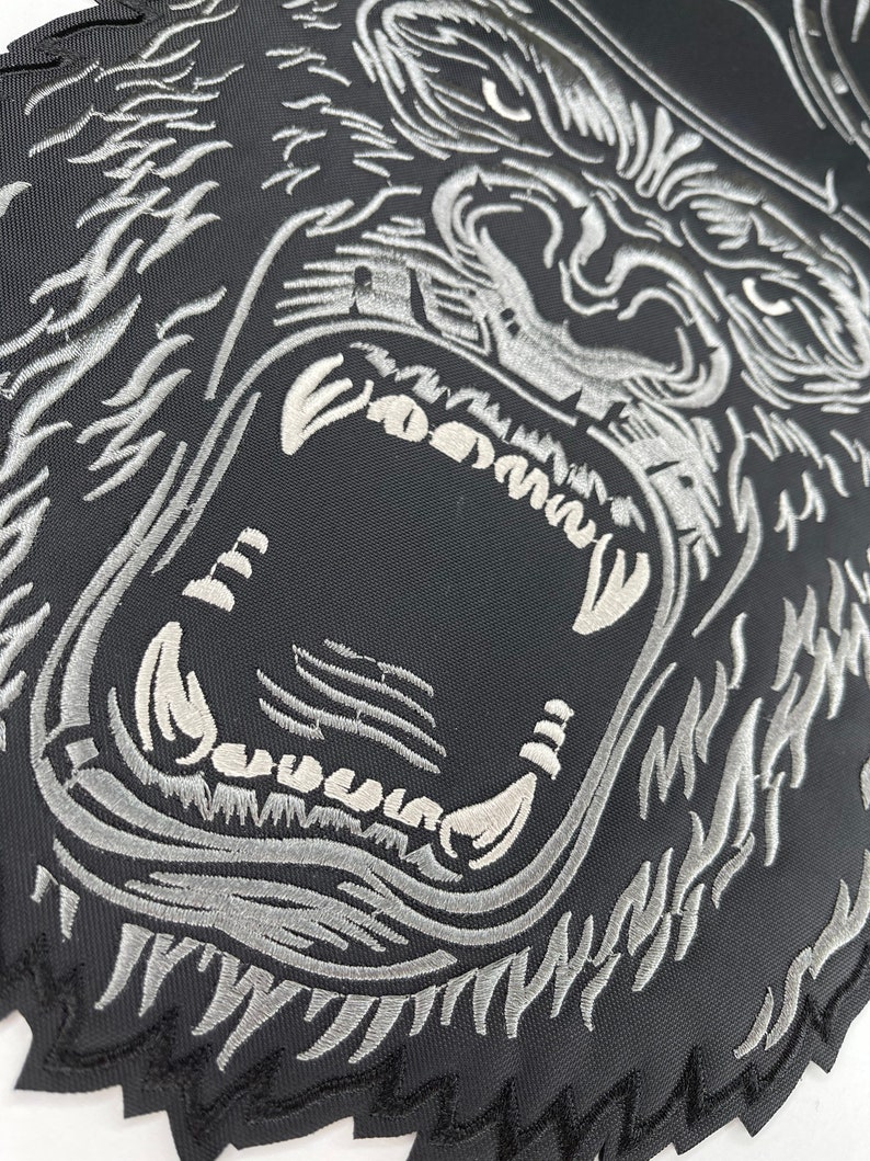 May include: A black and silver embroidered patch of a gorilla's face with a fierce expression. The gorilla's teeth are bared and its eyes are wide open.