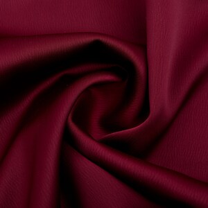 Puede incluir: Primer plano de una tela burdeos intenso con una superficie sutilmente texturizada. El material est&aacute; drapeado y doblado, creando sombras y reflejos que enfatizan su apariencia lujosa. El color rico y la textura suave sugieren un textil de alta calidad.