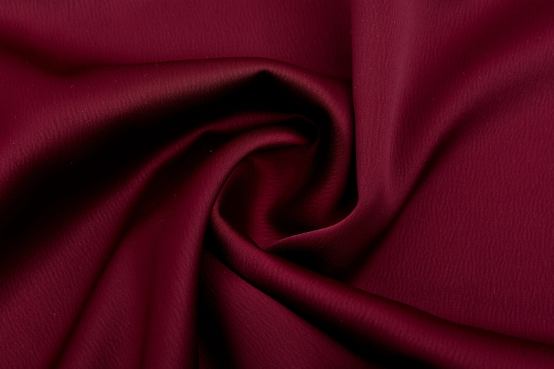 Puede incluir: Primer plano de una tela burdeos intenso con una superficie sutilmente texturizada. El material est&aacute; drapeado y doblado, creando sombras y reflejos que enfatizan su apariencia lujosa. El color rico y la textura suave sugieren un textil de alta calidad.