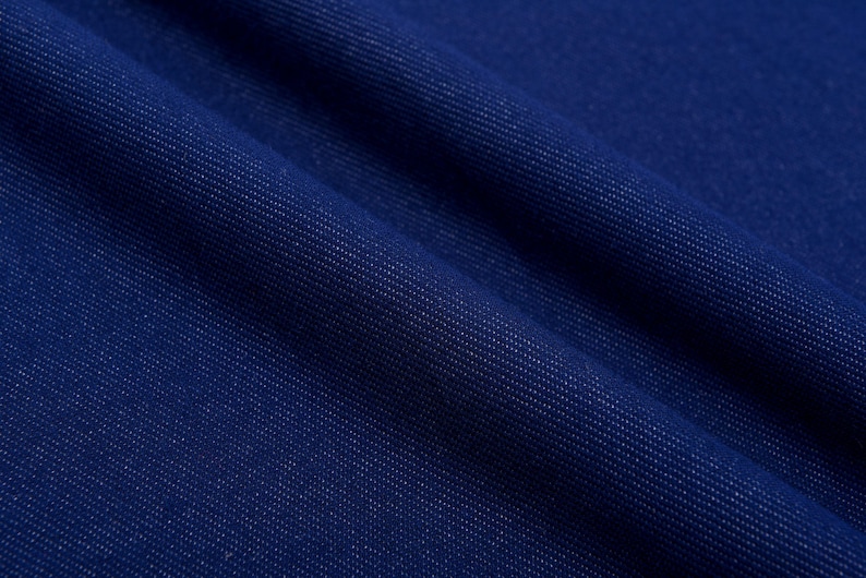 May include: Close-up of a deep blue denim fabric with a diagonal weave pattern. The fabric has a slightly textured surface and is folded, creating shadows and highlights. The color is a rich, saturated blue.