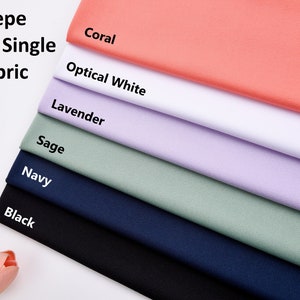 Pode incluir: Close-up de uma pilha de tecido de malha de crepe macio e el&aacute;stico em v&aacute;rias cores, incluindo coral, branco &oacute;ptico, lavanda, s&aacute;lvia, azul marinho e preto. O tecido &eacute; certificado com a certifica&ccedil;&atilde;o Oeko-Tex Standard 100.