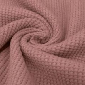 May include: Close-up of a dusty rose waffle knit fabric, tightly swirled to show texture. The fabric has a repeating grid pattern, creating a textured surface. The color is a muted pink, with subtle variations in tone due to the folds.
