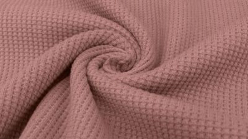 May include: Close-up of a dusty rose waffle knit fabric, tightly swirled to show texture. The fabric has a repeating grid pattern, creating a textured surface. The color is a muted pink, with subtle variations in tone due to the folds.