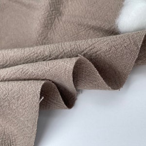 May include: Close-up of a brown linen fabric with a textured weave. The fabric is folded and draped, showcasing its soft and natural texture.