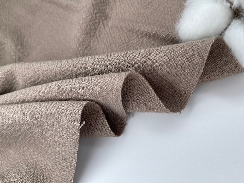 May include: Close-up of a brown linen fabric with a textured weave. The fabric is folded and draped, showcasing its soft and natural texture.