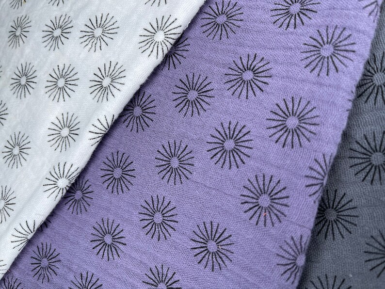 May include: Three fabric swatches with a repeating pattern of black sunburst designs. The swatches are white, light purple, and dark gray.