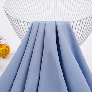 May include: A light blue fabric swatch draped over a white wire basket. The fabric has a smooth, slightly textured surface.