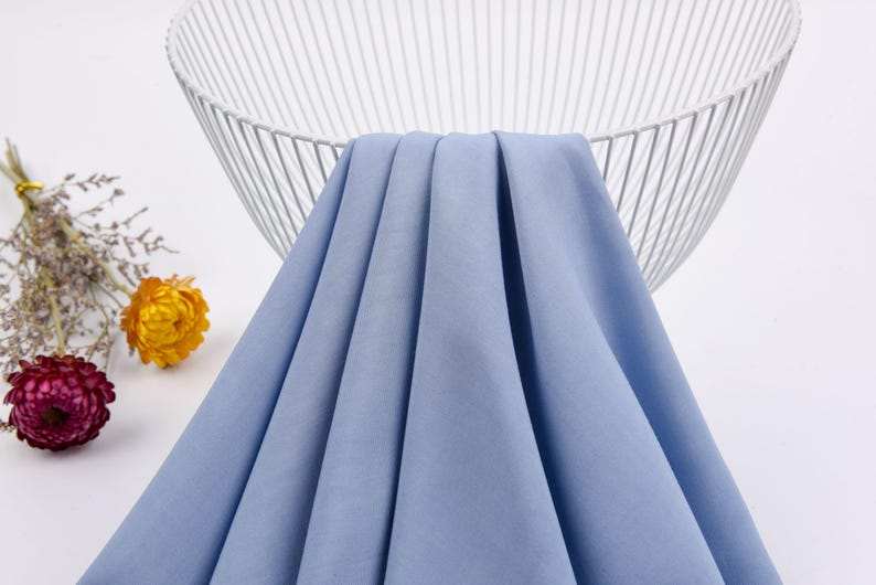 May include: A light blue fabric swatch draped over a white wire basket. The fabric has a smooth, slightly textured surface.