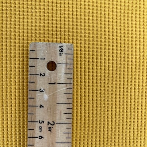 May include: Mustard yellow waffle knit fabric with a wooden ruler showing measurements in inches. The ruler is marked with 1/8 inch increments and shows a 2-inch measurement. The fabric has a textured, grid-like pattern.