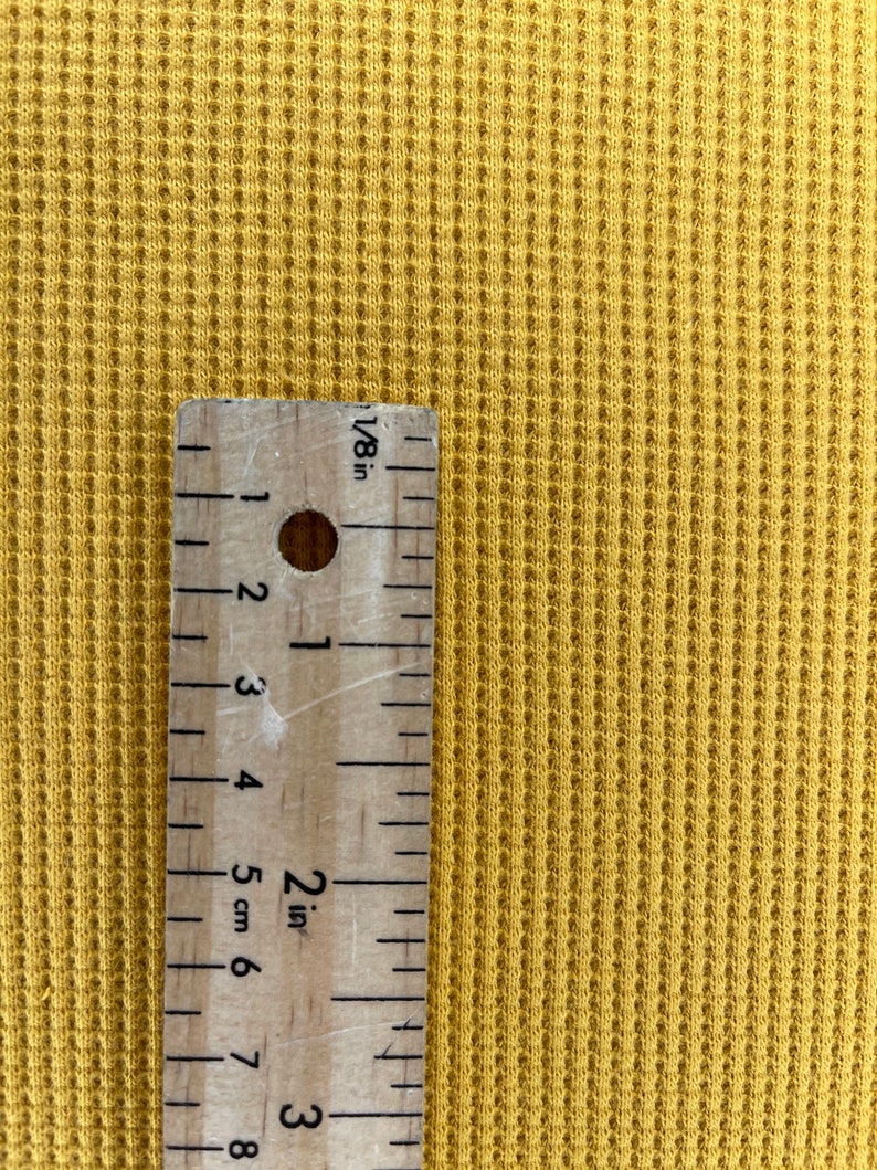 May include: Mustard yellow waffle knit fabric with a wooden ruler showing measurements in inches. The ruler is marked with 1/8 inch increments and shows a 2-inch measurement. The fabric has a textured, grid-like pattern.