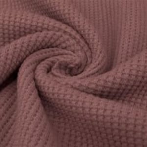 May include: Close-up of a mauve-colored waffle knit fabric. The fabric is tightly woven with a textured, grid-like pattern. The material is gathered and swirled, showing the texture and color variations. The fabric appears soft and pliable.