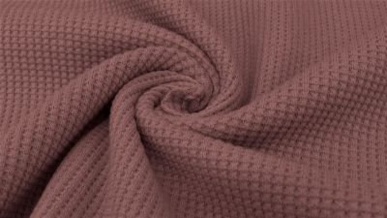 May include: Close-up of a mauve-colored waffle knit fabric. The fabric is tightly woven with a textured, grid-like pattern. The material is gathered and swirled, showing the texture and color variations. The fabric appears soft and pliable.