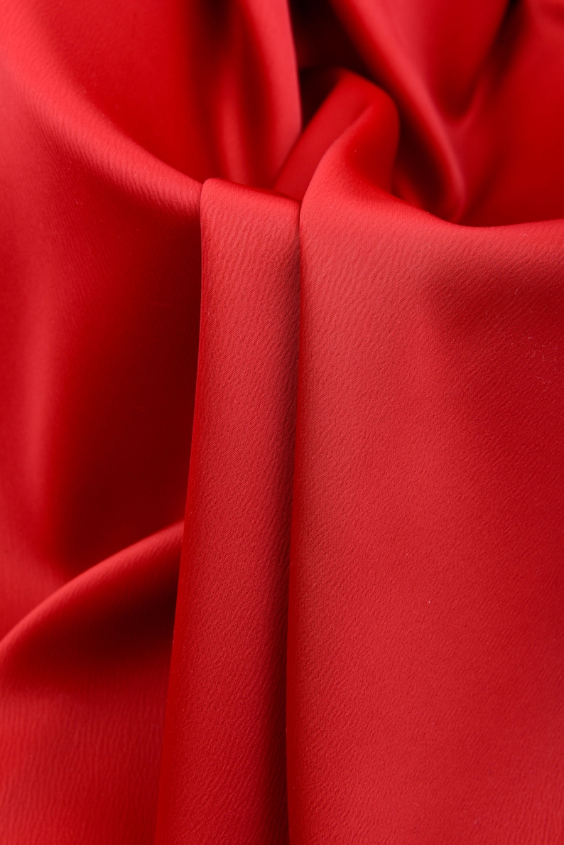 Puede incluir: Primer plano de tela roja vibrante con superficie texturizada. La tela est&aacute; drapeada y doblada, creando sombras y reflejos que enfatizan su textura suave. El efecto general es rico y lujoso.