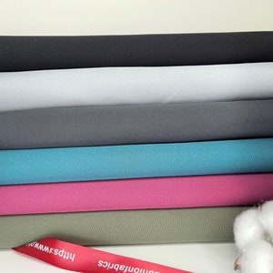 K&ouml;nnte beinhalten: Ein Stapel Stoffrollen in verschiedenen Unifarben, darunter Schwarz, Grau, T&uuml;rkis und Pink. Ein rotes Band mit wei&szlig;em Text ist unten sichtbar. Ein Zweig Baumwolle befindet sich im Hintergrund.