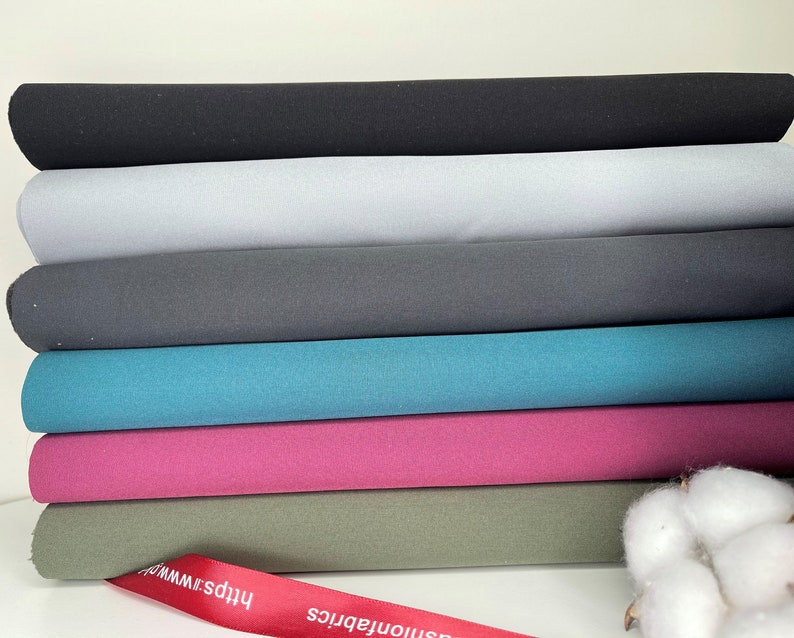 K&ouml;nnte beinhalten: Ein Stapel Stoffrollen in verschiedenen Unifarben, darunter Schwarz, Grau, T&uuml;rkis und Pink. Ein rotes Band mit wei&szlig;em Text ist unten sichtbar. Ein Zweig Baumwolle befindet sich im Hintergrund.