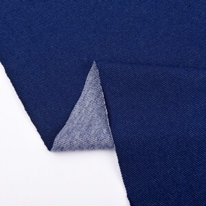 May include: Close-up of a swatch of dark blue denim fabric. The fabric is folded over, revealing the lighter, gray underside. The denim has a textured appearance and is set against a white background.