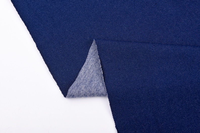 May include: Close-up of a swatch of dark blue denim fabric. The fabric is folded over, revealing the lighter, gray underside. The denim has a textured appearance and is set against a white background.