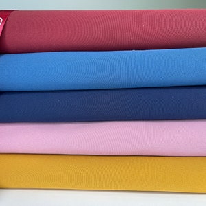 K&ouml;nnte beinhalten: Ein Stapel bunter Stoffrollen in den Farbt&ouml;nen Burgunderrot, Blau, Marine, Rosa und Senfgelb. Ein rotes Band mit dem Text "gkfashionsfabrics" ist &uuml;ber den Stoff drapiert. Der Stoff scheint ein weiches Strickmaterial zu sein.