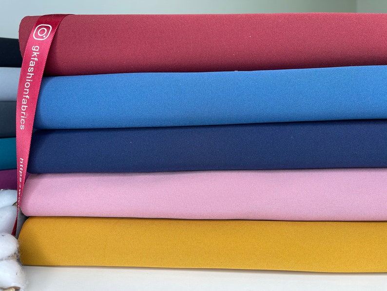 K&ouml;nnte beinhalten: Ein Stapel bunter Stoffrollen in den Farbt&ouml;nen Burgunderrot, Blau, Marine, Rosa und Senfgelb. Ein rotes Band mit dem Text "gkfashionsfabrics" ist &uuml;ber den Stoff drapiert. Der Stoff scheint ein weiches Strickmaterial zu sein.