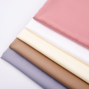 Puede incluir: Vista cenital de una colecci&oacute;n de muestras de tela dobladas en varios colores. Los colores incluyen tonos de rosa, blanco, crema, marr&oacute;n y lavanda. Las telas est&aacute;n dispuestas diagonalmente sobre un fondo blanco, mostrando sus texturas.