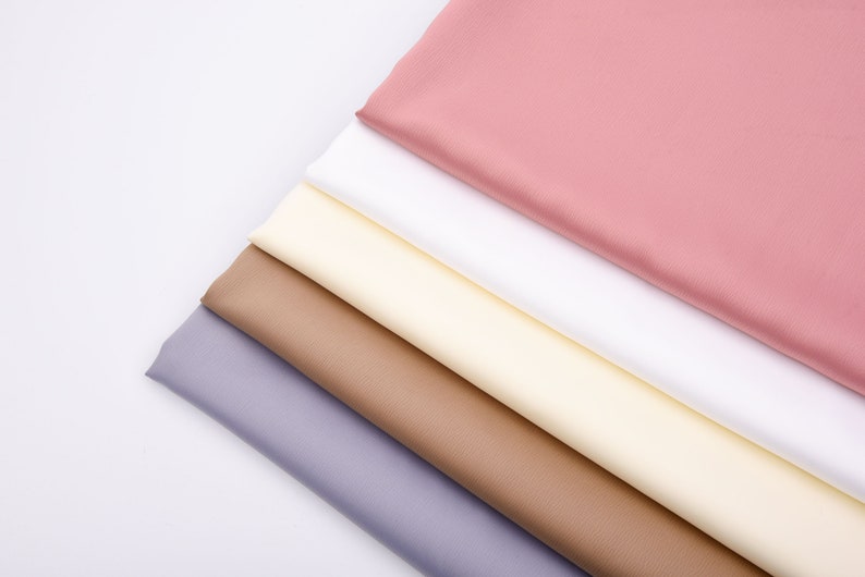 Puede incluir: Vista cenital de una colecci&oacute;n de muestras de tela dobladas en varios colores. Los colores incluyen tonos de rosa, blanco, crema, marr&oacute;n y lavanda. Las telas est&aacute;n dispuestas diagonalmente sobre un fondo blanco, mostrando sus texturas.