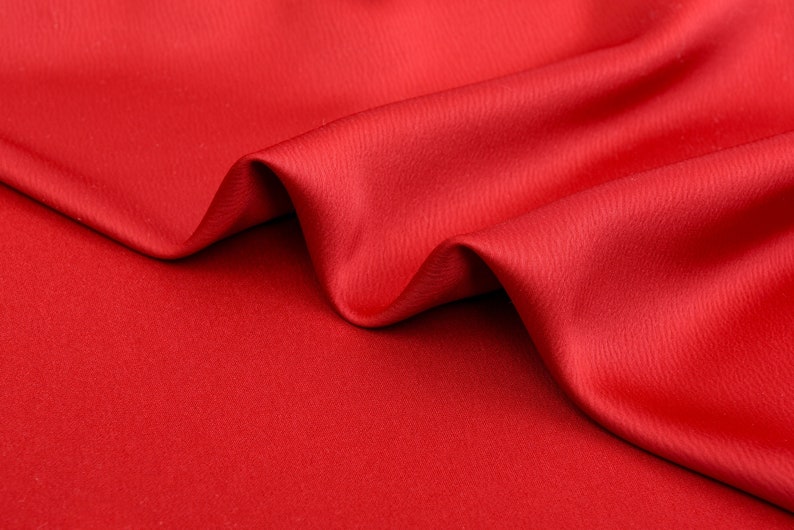 Puede incluir: Primer plano de una tela roja vibrante con pliegues suaves y un brillo sutil. El material parece liso y lujoso, con una ligera textura visible a la luz. La tela probablemente se utiliza para ropa o decoraci&oacute;n del hogar.