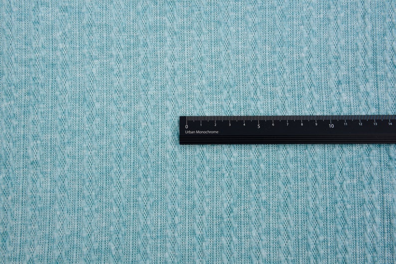 May include: A close-up of a teal textured fabric with a black ruler labeled "Urban Monochrome" placed on top. The ruler is marked in centimeters from 0 to 14.