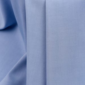 May include: Close-up of a light blue fabric with a smooth texture. The fabric is folded in a way that creates a series of vertical lines.